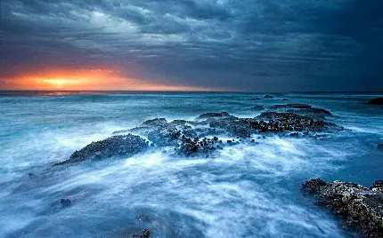 عکس برخورد امواج آبی اقیانوس به صخره ها با تابش نافذ و جالب خورشید در حال غروب با کیفیت اصلی