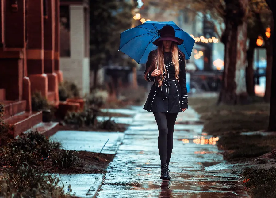 تصویر دلنشین از دختر باکلاس با کلاهی زیبا روی سرش و چتری آبی رنگ در باران  واقعی و کنار خانە‌های خوشنما