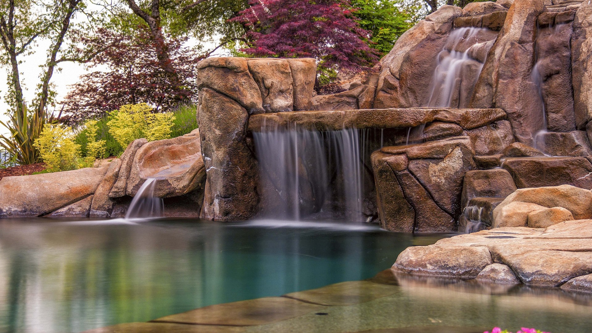 صفحه زمینه آبشار زلال و بهشتی با احاطه گیاهان بهاری برای دسکتاپ