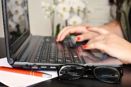 لپ تاپ و عینک طبی و دست های نویسنده زن با لاک نارنجی